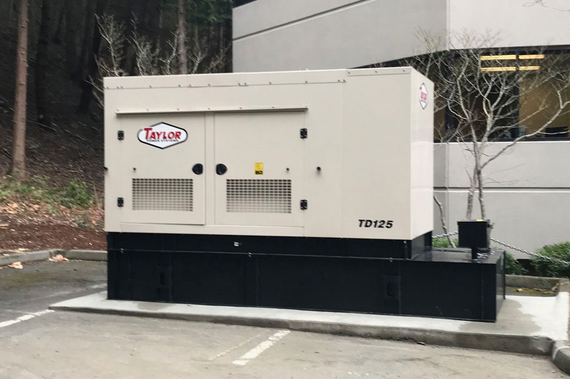A large beige and black Taylor TD125 generator from D2 Energy sits on a concrete platform outside a commercial building, near a parking lot and trees.