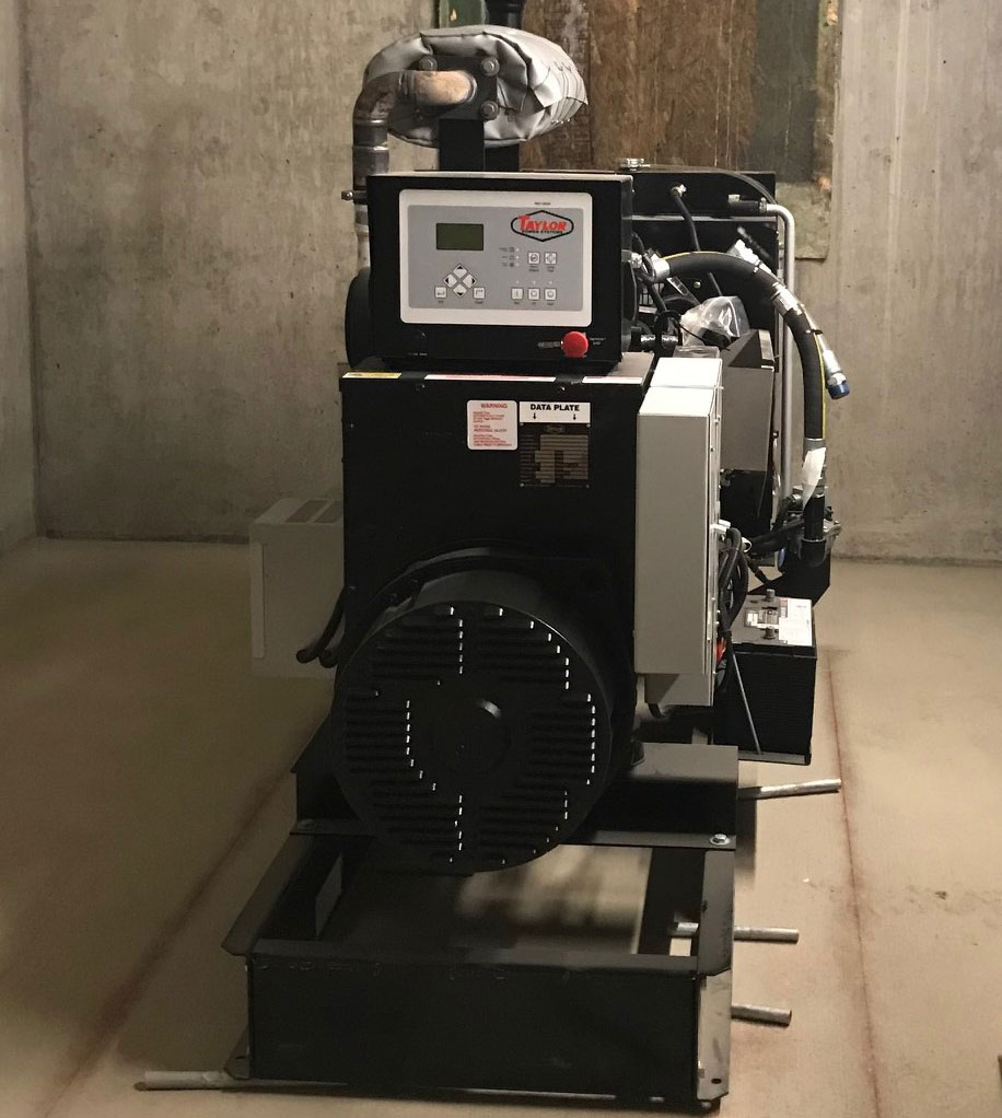 A D2 Energy commercial backup generator stands in a concrete-walled room. The generator features a control panel with buttons and a display screen, ventilation fans, and attached cables and pipes, all supported by a sturdy metal frame.
