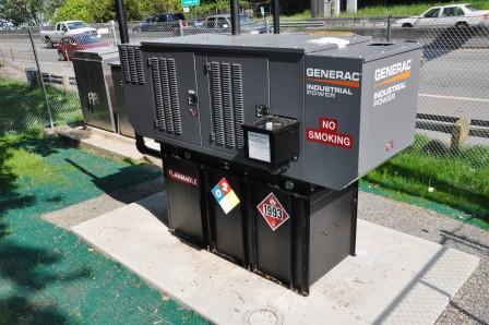 A large Generac industrial power generator sits on black fuel tanks outdoors, near a fence and road. Safety signs and a “No Smoking” notice are visible, highlighting the importance of professional generator services for safe operation.