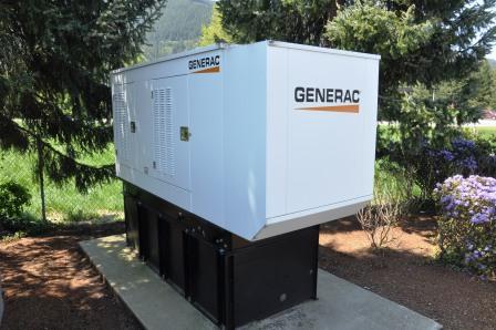 A large white Generac generator sits on a concrete pad outdoors, surrounded by trees and shrubs with mountains in the background—ideal for those seeking Yakima Generator Repair or maintenance in rugged outdoor settings.