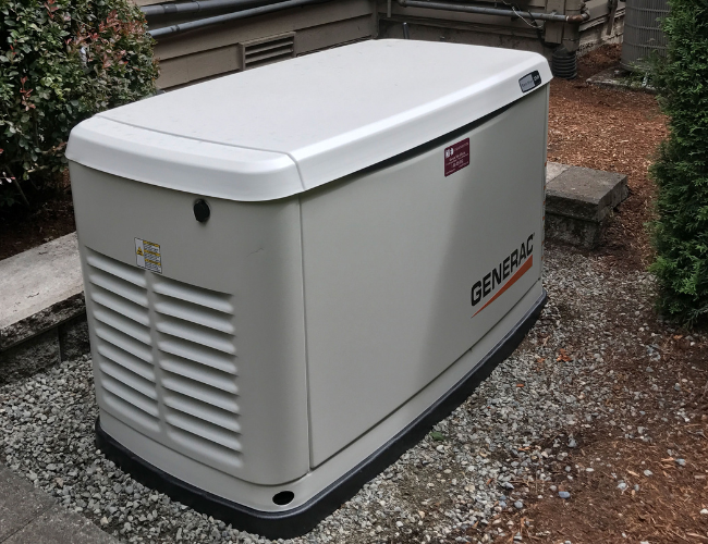 A large beige Generac standby generator from D2 Energy sits on a gravel bed outside a building, surrounded by shrubs, with ventilation slats visible on one side and power equipment in the background.