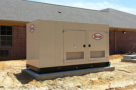 A large beige Taylor-branded generator sits on a concrete slab outside a brick building with windows and a shingled roof, on a patch of bare dirt—powered by reliable D2 Energy solutions.