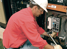 A man wearing a white hard hat, safety glasses, and red shirt is using tools to work on electrical equipment inside an open panel outdoors as part of Bellevue Generator Repair services.