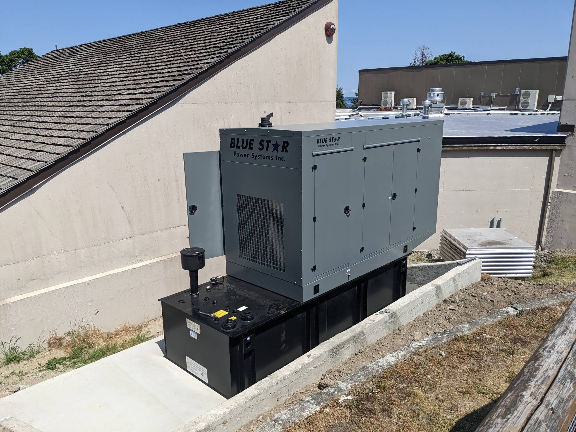 A large Blue Star Power Systems generator, maintained by a trusted generator repair company, is installed on a concrete pad outside a building. Nearby are HVAC units on the roof and stacks of white construction materials.